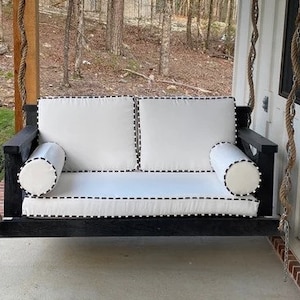 May include: A white porch swing with black trim and rope hanging from the ceiling. The swing has two white pillows with black stitching.