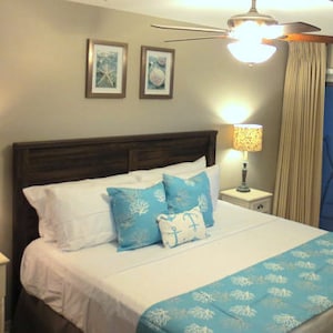 May include: A bedroom with a king-sized bed with white bedding and blue and white patterned pillows. The bed has a blue and white patterned bed runner. There are two nightstands with lamps on either side of the bed. The room has a ceiling fan and a sliding glass door leading to a balcony.