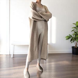 May include: A beige, longline cardigan with a matching vest top and leggings. The outfit is completed with matching slip-on shoes. The ensemble is neutral-toned and appears to be made of a soft, knit material. The cardigan has long sleeves and a relaxed fit.