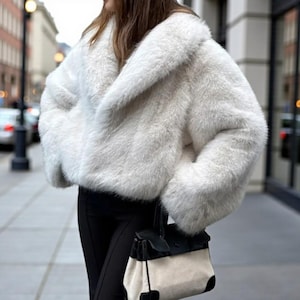 Könnte beinhalten: Eine weiße Kunstfelljacke mit breitem Kragen. Die Jacke wird mit einer schwarzen Hose und einer schwarz-weißen Handtasche kombiniert. Die Jacke hat eine weiche, flauschige Textur und eine verkürzte Länge, für einen stilvollen Look.