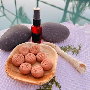 May include: A red spray bottle with a black label and cap, next to a wooden bowl filled with small, round, pinkish-brown objects, each with a symbol. Two gray stones and a piece of driftwood are also present.