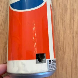 May include: A cylindrical object with a red, white, and blue design, reminiscent of a soda can. The object has a rectangular port and the word "ENTER" is visible. The object is on a wooden surface.