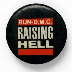 May include: A black round button with the text "RUN-D.M.C. RAISING HELL" in white letters on a red background. The button is a vintage item, likely from the 1980s, and is a collectible item for fans of the hip-hop group.