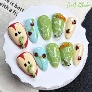 Könnte beinhalten: Ein Set aus zehn künstlichen Fingernägeln mit Naturmotiven. Die Designs umfassen rote Apfelscheiben mit grünen Würmern, hellblaue Nägel mit Marienkäfern, grüne Blätter mit Wassertropfen sowie cremefarbene Nägel mit Marienkäfern und orangefarbenen Raupen.