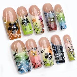 May include: A set of ten artificial nails with a nude base and colorful designs. Each nail features a French tip, floral accents, and sparkling rhinestones. The designs include gradients of green, orange, pink, blue, and black.