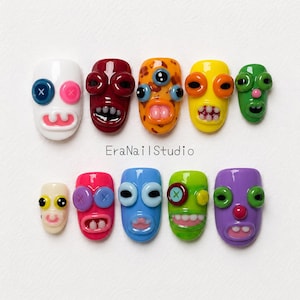 May include: A set of ten press-on nails featuring colorful, glossy monster faces with raised button eyes, protruding mouths, and teeth. The text EraNailStudio is printed in the center. Each nail displays a unique combination of colors and facial features.