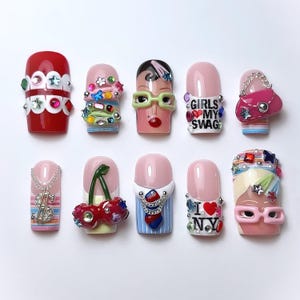 May include: A set of ten press-on nails featuring diverse 3D designs including a red pattern, a face with glasses, a pink handbag, a dollar sign charm, cherries, a necktie, and text reading GIRLS MY SWAG and I LOVE NY with colorful rhinestones.