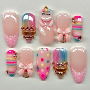 May include: A set of ten press-on nails featuring various designs including pink polka dots with white scalloped edges, iridescent gradients with gold and pink rhinestones, a 3D unicorn figure, pink bows, and multi-colored horizontal stripes.