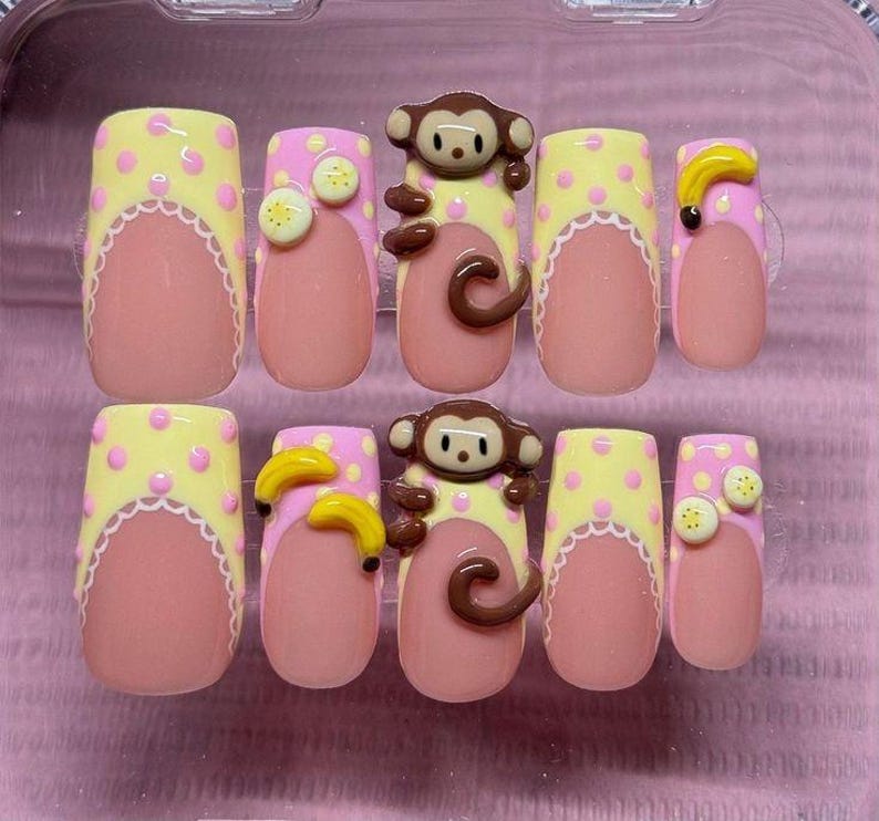 May include: A set of press-on nails with a playful design. The nails feature a light pink base with a scalloped white edge, topped with yellow polka dots. Some nails have 3D monkey and banana embellishments.