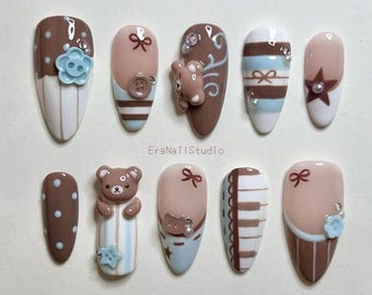 Op de afbeelding: Een set van tien amandelvormige press-on nagels met een kleurenpalet van bruin, wit en lichtblauw. De ontwerpen bevatten een 3D-teddybeer, kleine knoopjes, witte stippen, horizontale strepen, een ster en pianotoetsen. De tekst EraNailStudio is zichtbaar.