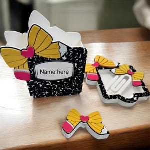 Può includere: Un organizer da scrivania, un portaclip e un abbellimento a forma di matita. L'organizer e il supporto presentano un design a quaderno di composizione in bianco e nero con un fiocco giallo e un accento di matita a cuore rosa. Il testo "Name here" è visibile.