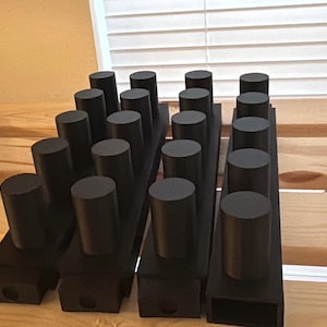 May include: Four rectangular black bases arranged in parallel, each featuring a row of four vertical black cylindrical pegs extending upward. The objects have a matte finish and a uniform design, with a small circular opening visible at the front base.