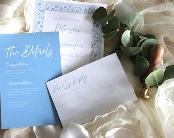 May include: Wedding invitation suite featuring a light blue card with the words "The Details" and a white script font. A matching RSVP card and invitation are also present, along with greenery and a wooden stamp.