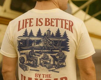 May include: Cream-colored t-shirt with a graphic design featuring an airplane, two figures, and the text "LIFE IS BETTER BY THE HANGAR" in red. The design evokes a vintage, outdoorsy theme.