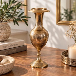 May include: A tall, polished brass vase featuring an intricate floral engraving on its bulbous body, a slender neck, and a flared rim. The metal surface displays horizontal etched bands and rests on a circular pedestal base with detailed carvings.