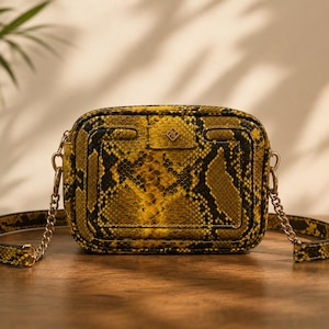 May include: A rectangular crossbody bag with a snakeskin pattern in shades of yellow and black. The bag has a gold chain strap and a small gold emblem on the front. The bag is sitting on a wooden surface.