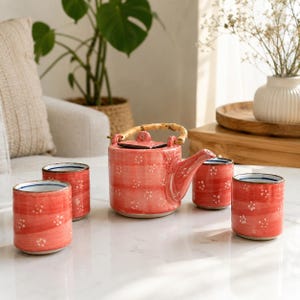 Puede incluir: Un juego de té de cerámica que incluye una tetera con asa de bambú curvada y tres tazas cilíndricas a juego. Las piezas tienen un acabado en esmalte rojo coral vibrante con pinceladas horizontales y pequeños motivos florales blancos.