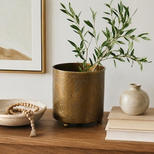 May include: A cylindrical brass planter with intricate embossed patterns and three small feet, containing a green leafy plant. Beside it sits a shallow stone bowl with wooden beads and a small beige ceramic vase resting on two stacked books.