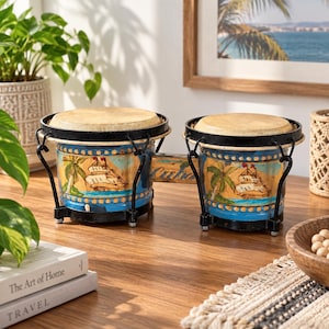 May include: A set of two bongo drums connected by a wooden bridge with the word Cuba painted on it. The drums feature a blue base with hand-painted illustrations of palm trees and sailing ships, topped with tan drumheads and black metal rims.
