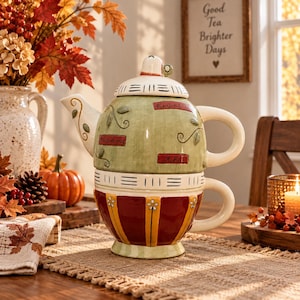 May include: A ceramic tea-for-one set featuring a stacked teapot and teacup. The teapot is sage green with black line details, leaf motifs, and red rectangular labels reading herbal and tisane. The teacup has red and yellow vertical stripes.