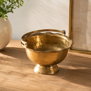 Puede incluir: Un cuenco de latón pulido con textura martillada, borde festoneado y asa articulada. El cuenco se apoya sobre una base circular de pedestal, mostrando un acabado metálico dorado con reflejos de luz visibles en su superficie curva.