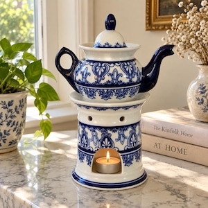 May include: A white ceramic teapot with a blue floral pattern and a matching lid, resting on a cylindrical ceramic warmer base. The base features a cutout for a small lit tea light candle, and both pieces are decorated with a consistent blue pattern.