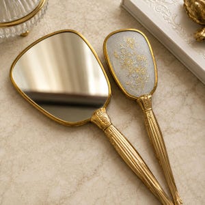 May include: A handheld mirror and a matching hairbrush with gold-toned metal handles featuring ornate, embossed detailing. The back of the hairbrush has a textured, light grey fabric inlay decorated with a delicate gold and silver floral pattern.
