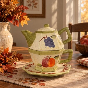 May include: A ceramic tea-for-one set featuring a teapot stacked on a teacup and saucer. The cream-coloured pieces have a speckled finish, green rims, and hand-painted fruit motifs including purple grapes on the teapot and an apple on the cup.