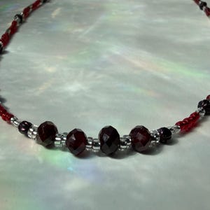 May include: A beaded necklace featuring faceted, dark red beads interspersed with smaller red and silver beads. The necklace has a delicate design, with a mix of textures and colors.