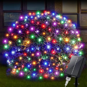 Puede incluir: Un arbusto vibrante cubierto de luces de cadena LED multicolores. Las luces son una mezcla de rojo, verde, azul y amarillo, creando una exhibición festiva. Un pequeño panel solar es visible a la derecha, lo que indica un sistema de iluminación alimentado por energía solar.