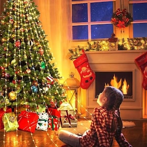 Puede incluir: Un árbol de Navidad decorado con luces y adornos, rodeado de regalos envueltos. Una chimenea con fuego, calcetines navideños y una ventana con vista al cielo nocturno completan la escena festiva.