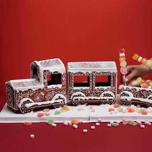 May include: A gingerbread train consisting of an engine, a passenger car, and a cargo car filled with colorful gummy candies. The brown gingerbread structures are decorated with white icing patterns and white icing snow on the roofs.