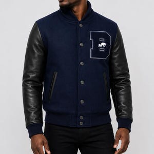Può includere: Una giacca varsity blu navy con maniche in pelle nera, colletto alla coreana e chiusura frontale con bottoni. Sul petto sinistro è presente una grande toppa bianca a forma di lettera B con un piccolo grafico di un elefante all'interno.