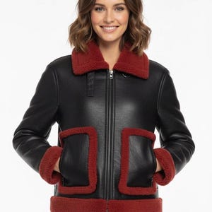 Puede incluir: Chaqueta de cuero negra con cuello, puños y ribetes de bolsillo de piel sintética roja. La chaqueta tiene cierre de cremallera frontal y dos bolsillos delanteros. Una prenda de abrigo elegante.
