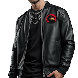 May include: Black faux leather bomber jacket with a red and black patch on the chest. The jacket has a silver zipper and a black ribbed collar and cuffs. The person is wearing a silver chain necklace and black pants.