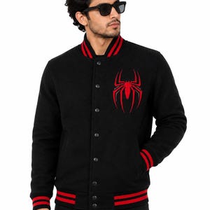 May include: A black varsity jacket featuring a red spider emblem on the left chest. The jacket has a snap-button front closure, a ribbed collar, cuffs, and hem, all accented with two horizontal red stripes. The fabric appears to be a wool-like material.