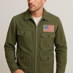 May include: Olive green jacket with a zipper closure, four front pockets, and an American flag patch on the chest. The jacket is worn over a cream-colored shirt and paired with blue jeans.