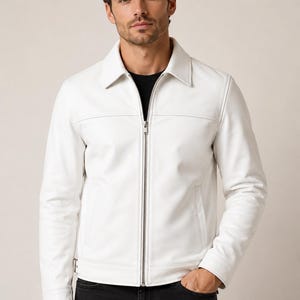 Op de afbeelding: Een witte leren jas met een kraag en een zilveren ritssluiting. De jas heeft twee voorzakken en wordt gedragen met een zwarte jeans. Klassieke stijl.