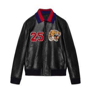 May include: Black leather jacket with a ribbed collar in blue and red. Features a red and white "25" patch and a tiger head patch. The jacket has a ribbed hem and cuffs.