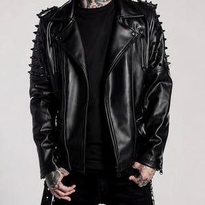 Puede incluir: Chaqueta de cuero negra estilo biker con cuello de solapa, cremallera asimétrica y detalles de tachuelas en hombros y mangas. La chaqueta tiene múltiples bolsillos con cremallera y cinturón.