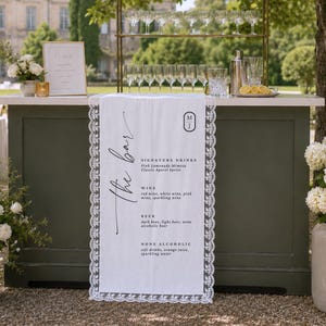 Puede incluir: Una barra portátil de color verde oscuro con un camino de mesa blanco que muestra el texto 'the bar' y un menú de bebidas, vino, cerveza y opciones sin alcohol. La barra incluye un estante de metal dorado con cristalería y una coctelera.