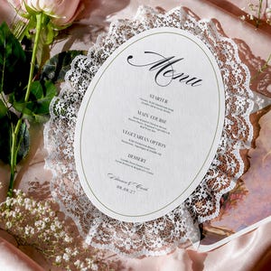May include: An oval-shaped paper menu with white lace trim, featuring the text Menu, Starter, Main Course, Vegetarian Option, Dessert, and the date 08.08.27. It is accompanied by a light pink rose and small white baby's breath flowers.