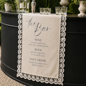 May include: A beige fabric table runner with intricate white lace trim, featuring the black printed text "the Bar" followed by a menu listing wine, beer, and soft drink options. The runner is draped over a curved black bar counter.