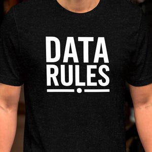 May include: Black t-shirt with the white text "DATA RULES" in a bold, sans-serif font. The text is centered and stacked, with a horizontal line and a dot beneath the word "RULES". The shirt is a short-sleeved crew neck.