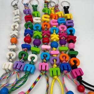 May include: A collection of colorful keychains with letter and bead accents. Each keychain features a silver keyring, followed by letter beads spelling out a word, and colorful beads. The keychains are strung on a variety of cords.