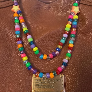 May include: A brown leather handbag with a beaded strap. The strap is made of colorful beads in various colors, including pink, orange, yellow, green, blue, and purple. The strap has a star-shaped charm. The bag has a gold-colored metal plate with text.