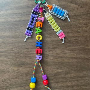 May include: A keychain featuring a green star-shaped carabiner with multiple beaded strands. One strand spells TEACHER in colorful letter beads. Other strands include a purple and black pattern, a yellow and pink pencil design, and a blue and silver grid.