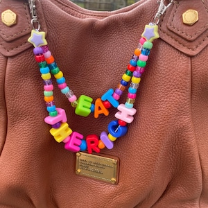 May include: A brown leather handbag with gold hardware is adorned with a beaded strap. The strap features colorful beads spelling out "EAT RICH" in bright, playful letters. A star-shaped charm adds a whimsical touch.