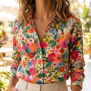 May include: A floral print button-down shirt with long sleeves, featuring a vibrant mix of pink, orange, blue, and yellow flowers on a cream background. Paired with beige linen pants. The shirt has a classic collar and is partially unbuttoned.