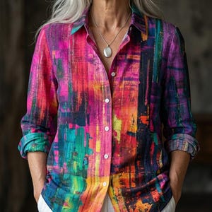 May include: A button-down shirt with a vibrant abstract design in shades of pink, purple, orange, and teal. The long-sleeved shirt has a collared neckline and white buttons. The shirt is paired with white pants.